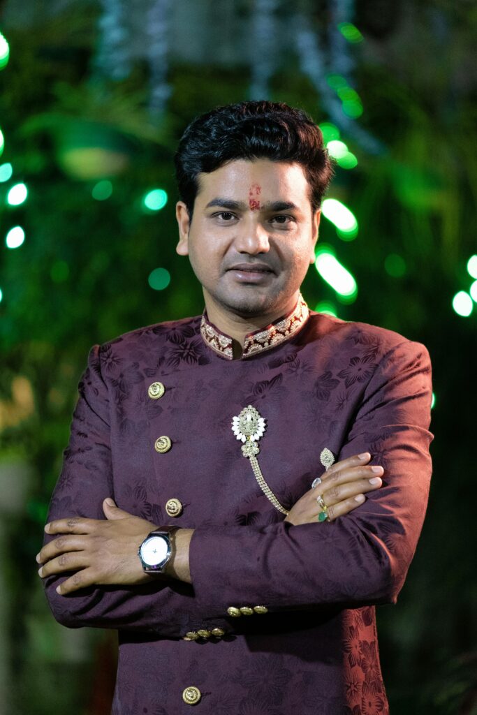 An Indian man in traditional attire with tilak, posing with arms crossed in an elegant setting.